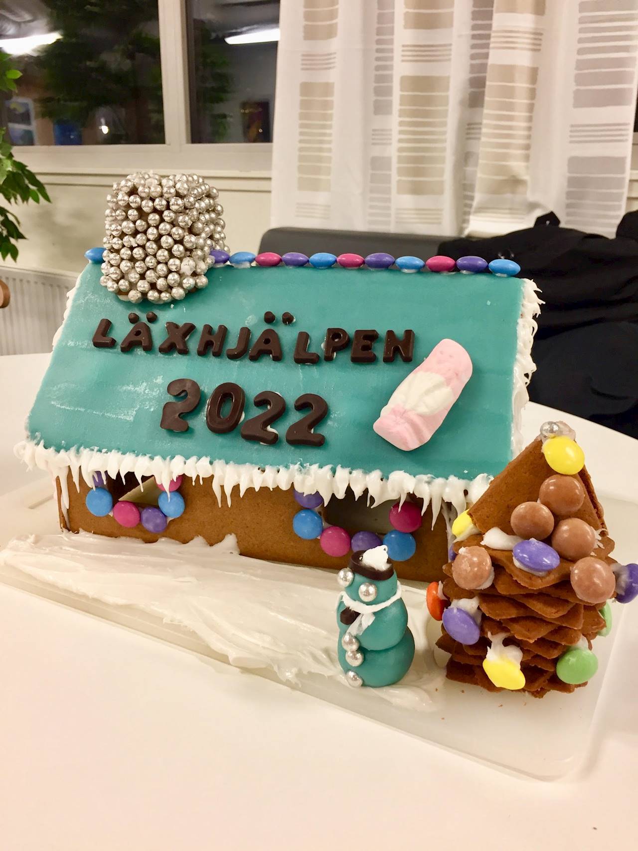 Pepparkakshus med ett tak i blå glasyr med en skumtomte på och en skorsten gjord av silverfärgade chokladkulor. På taket står det "Läxhjälpen 2022" och framför huset står en julgran av pepparkaka och en snögubbe gjord av glasyr.