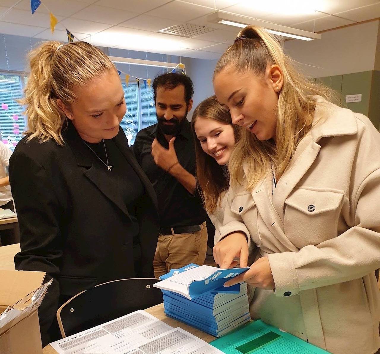 Tre läxhjälpare och en coach bläddrar i Läxhjälpens målböcker som ligger staplade på varandra på ett bord i ett upplyst klassrum