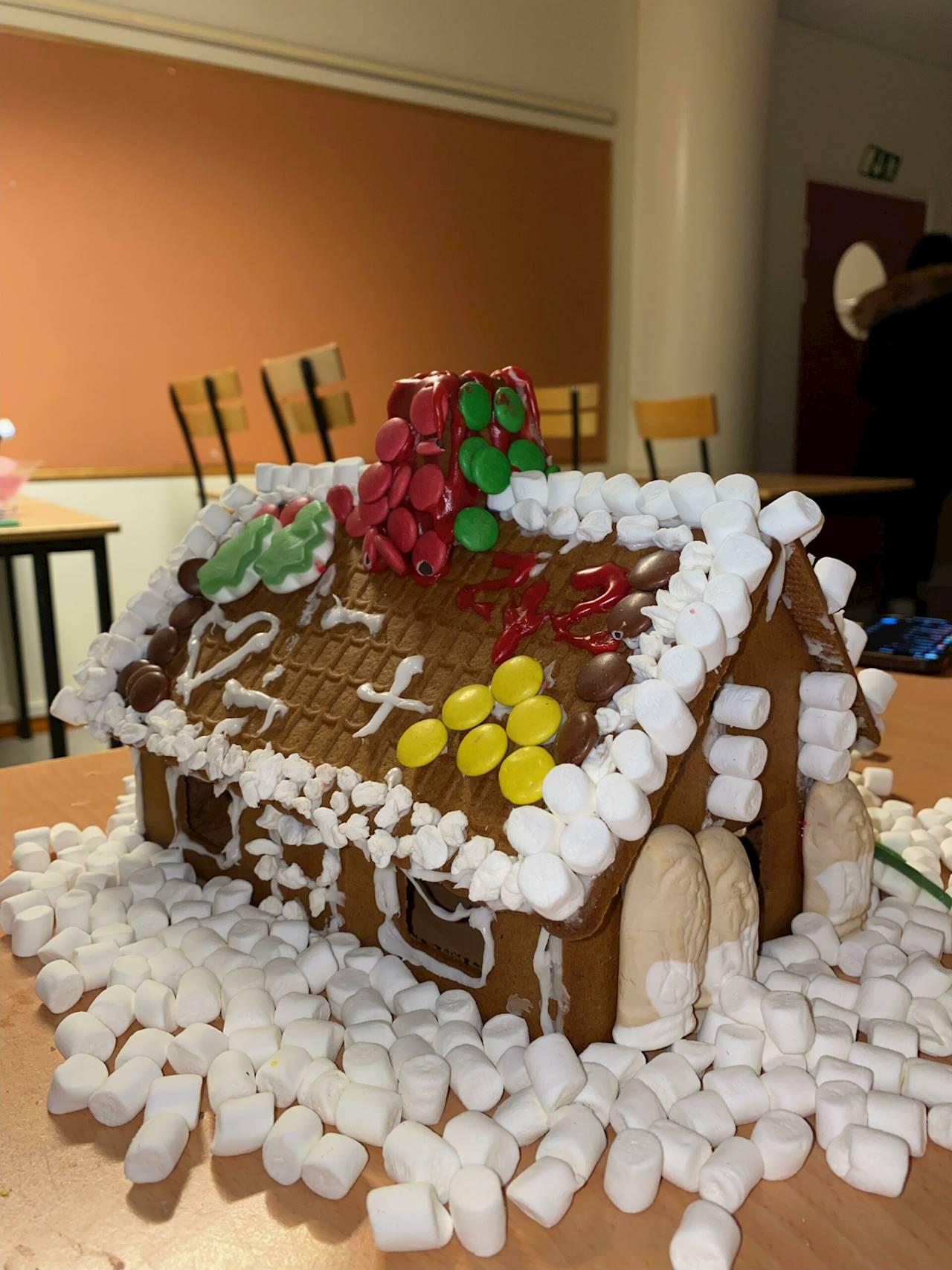 7. Nybyggeskolan MO1 (Västerås). Bilden föreställer ett pepparkakshus med marshmallows runt kanterna på taket. Taket är beklätt med ett hjärta i vit glasyr, ett kors i vit glasyr samt gula nonstop-choklad i form av ett A. Skorstenen är beklädd med gröna och röda nonstop-choklad. Fönstren på pepparkakshuset har vit glasyr runt kanterna. Nedanför huset, på marken, finns där marshmallows föreställande snö. Mot väggen till pepparkakshuset står tre skumtomtar.