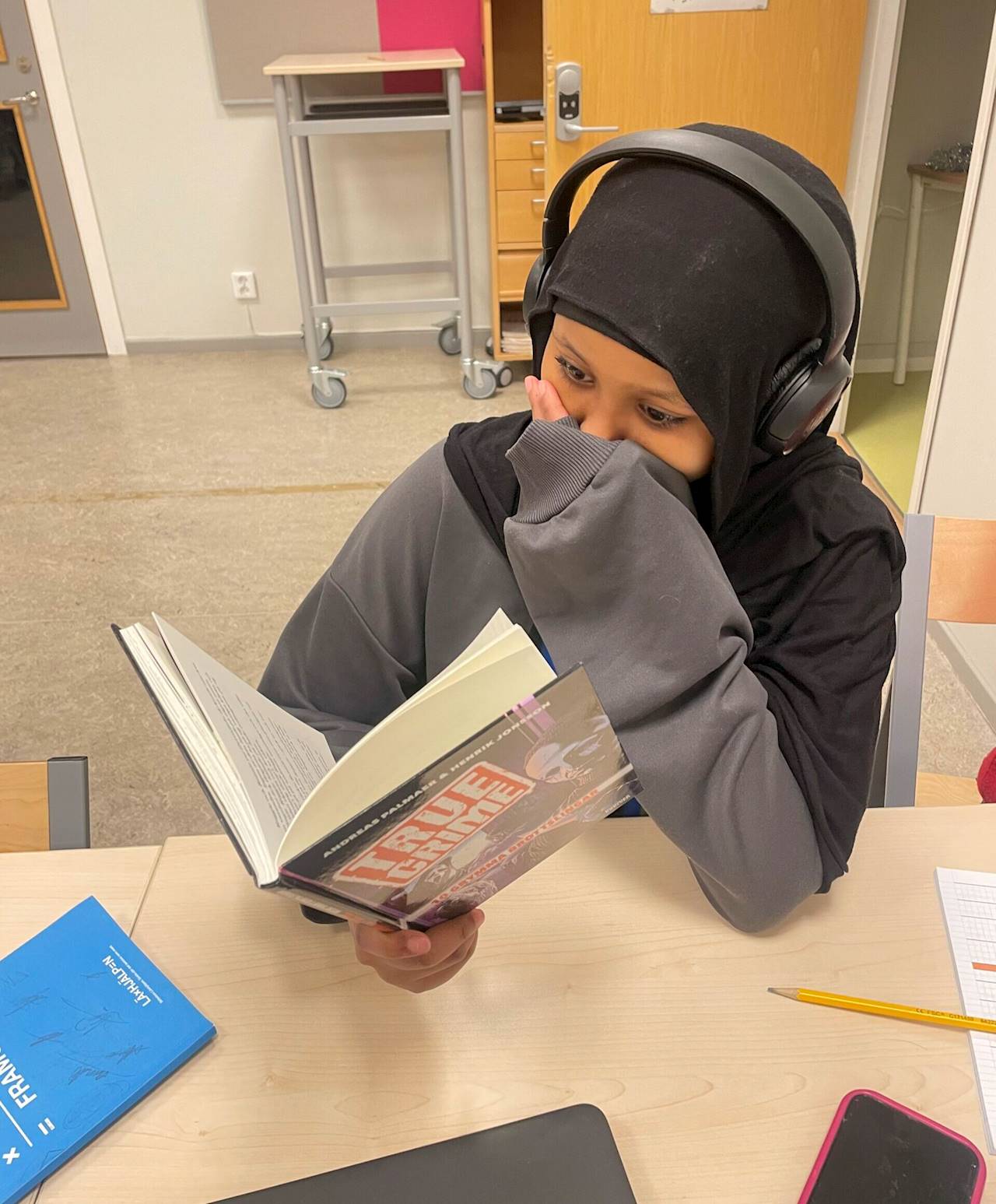En elev sitter vid en skolbänk i ett klassrum och läser i en bok. I ena handen håller hon i boken och andra handen håller hon för munnen. Hon har hörlurar på sig och framför henne, på bänken, ligger Läxhjälpens blåa målbok.