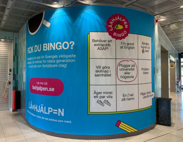 Väggvepa med Läxhjälpens kampanj "Fick du bingo?" på Flemingsbergs pendeltågsstation. Bakgrundsfärgen på väggvepan är blå med vit text och färgglada kaffe- och pennsymboler.