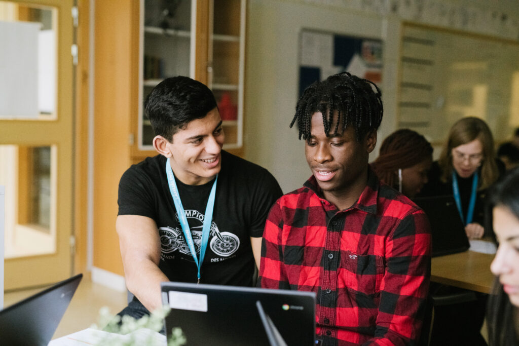 En läxhjälpare och en elev sitter bredvid varandra i ett klassrum och tittar gemensamt på en datorskärm framför dem. Läxhjälparen tittar på eleven och pekar på datorn och båda ler