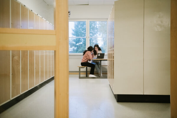 Två elever sitter vi ett bord i en korridor långt borta i bakgrunden. I förgrunden syns flera elevskåp längs med korridoren