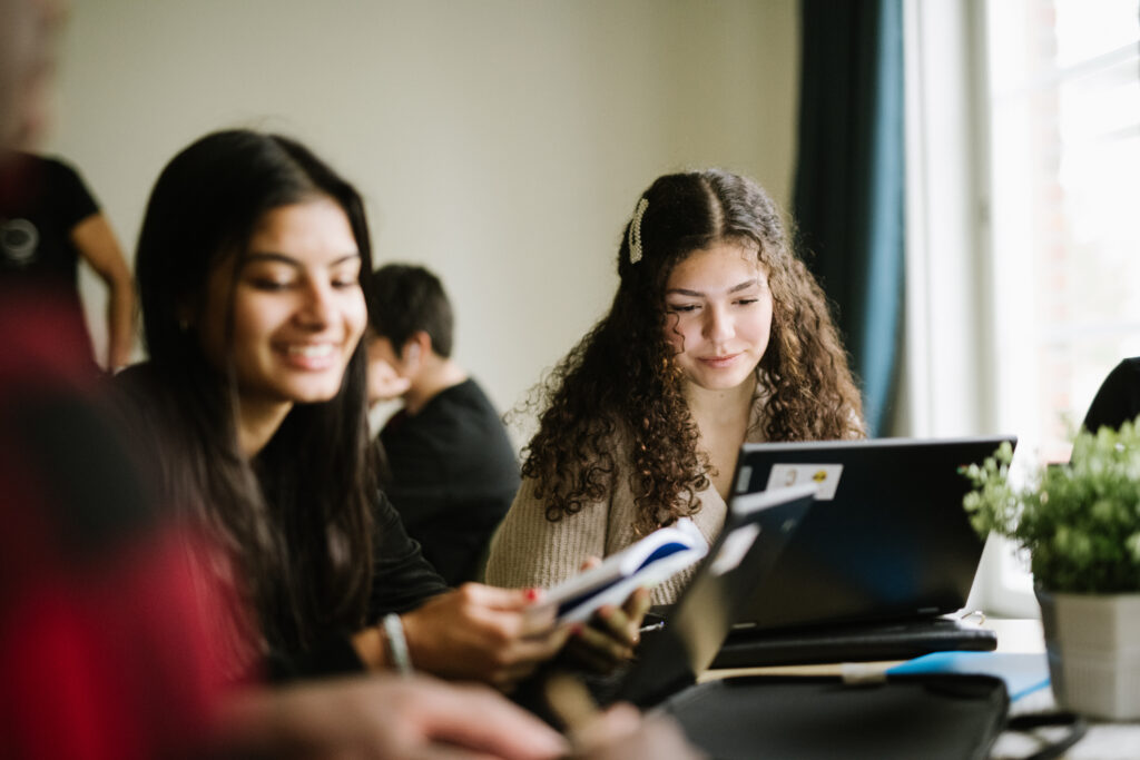 Två elever sitter vid ett bord i ett klassrum med dator och böcker framför sig och ler