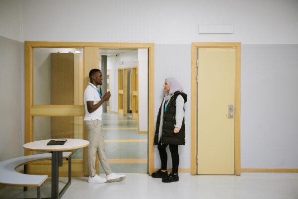 Två läxhjälpare står i en skolkorridor och diskuterar.