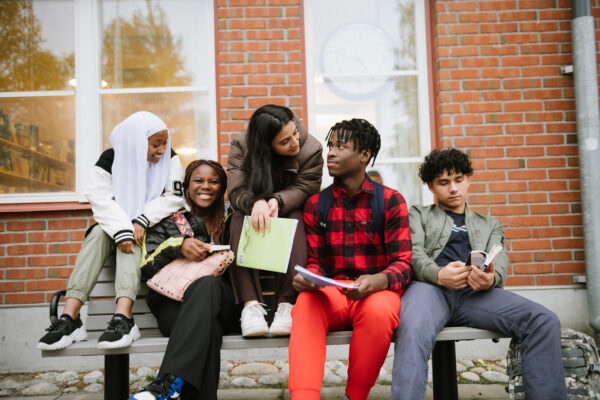 Fem elever sitter på en bänk på skolgården utan för en skolbyggnad. En elev tittar på sin kompis som skrattar, de två eleverna i mitten tittar på varandra och den femte eleven, till höger, tittar ner i sina läxor.