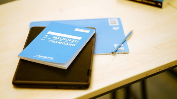 Bild på en laptop, ett anteckningsblock, en blå bok och en penna som ligger på ett skrivbord. På den blå boken står det "ditt namn x Målboken = framgång" med Läxhjälpens logotyp på.