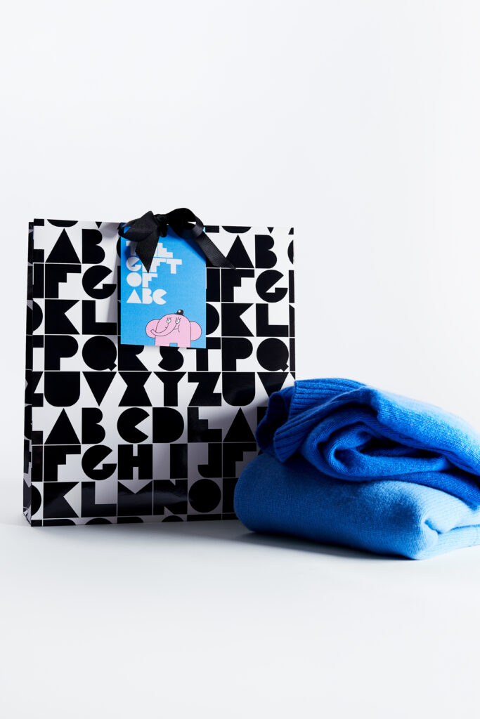 Bild på en svartvit presentpåse med ett mönster gjort av bokstäver. På kortet syns ett blått kort med en illustration av Owe Gustafson. Bredvid presentpåsen ligger ett blått klädesplagg.