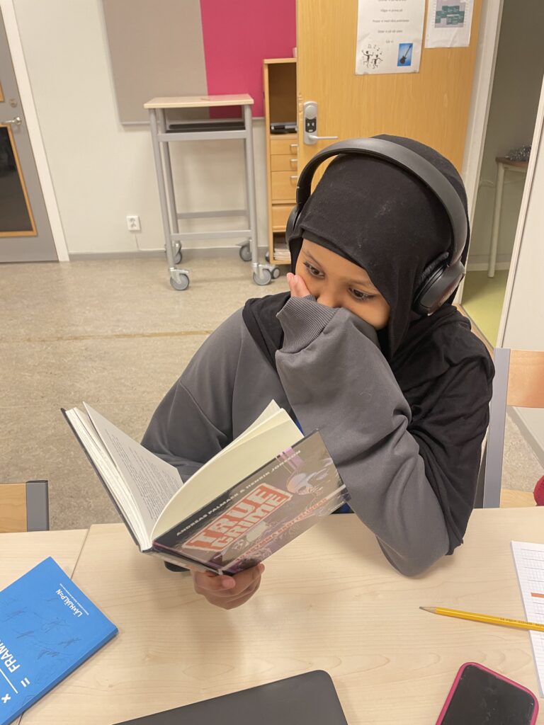 En elev sitter vid en skolbänk i ett klassrum och läser i en bok. I ena handen håller hon i boken och andra handen håller hon för munnen. Hon har hörlurar på sig och framför henne, på bänken, ligger Läxhjälpens blåa målbok.