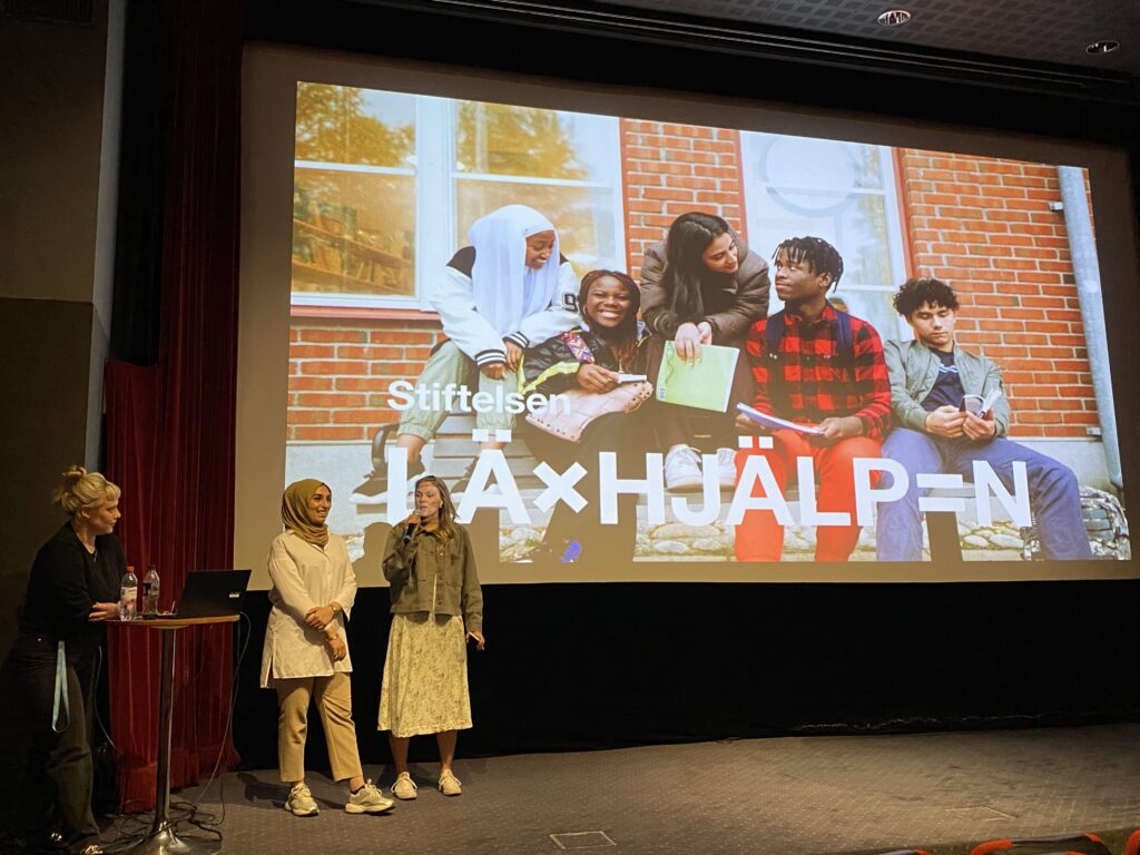 Tre personer står uppe på en biografscen och presenterar framför en skärm. På skärmen syns en presentationsbild på en grupp elever och Läxhjälpens logotyp i vitt