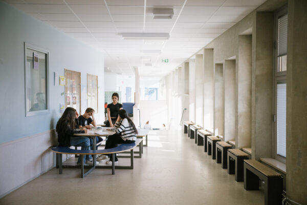 Fotografi av en skolkorridor med bänkar längs fönstren på högersidan och ett bord längs väggen på vänstersidan. Vid bordet sitter tre elever och studerar och en fjärde elev står upp och håller sina skolböcker i händerna.
