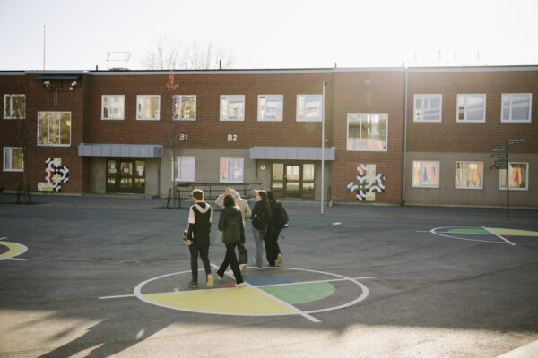Fotografi av en skolgård i eftermiddagssol där ryggarna på fem elever som går över skolgården syns. I bakgrunden ligger en skolbyggnad i skugga.