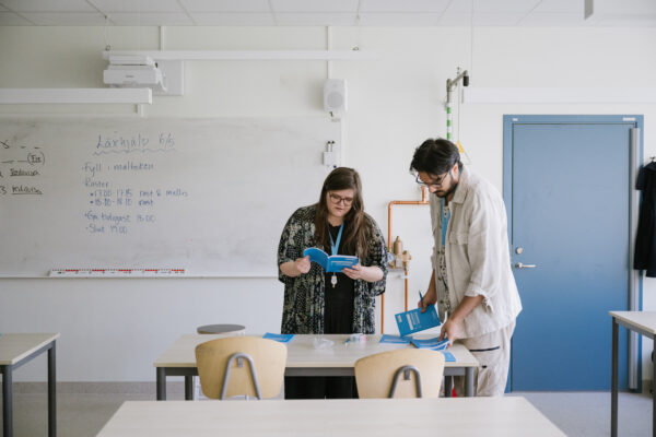 Fotografi av två läxhjälpare som står i ett klassrum och bläddrar bland Läxhjälpens blå målböcker. I bakgrunden syns en whiteboard-tavla där det står "Läxhjälp 6/5" och schemat för dagen.