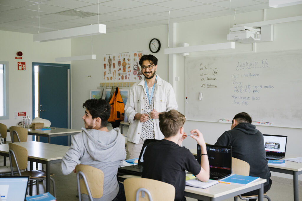 En leende ung man står med ryggen mot en whiteboard-tavla i ett klassrum. Tre elever sitter framför honom.